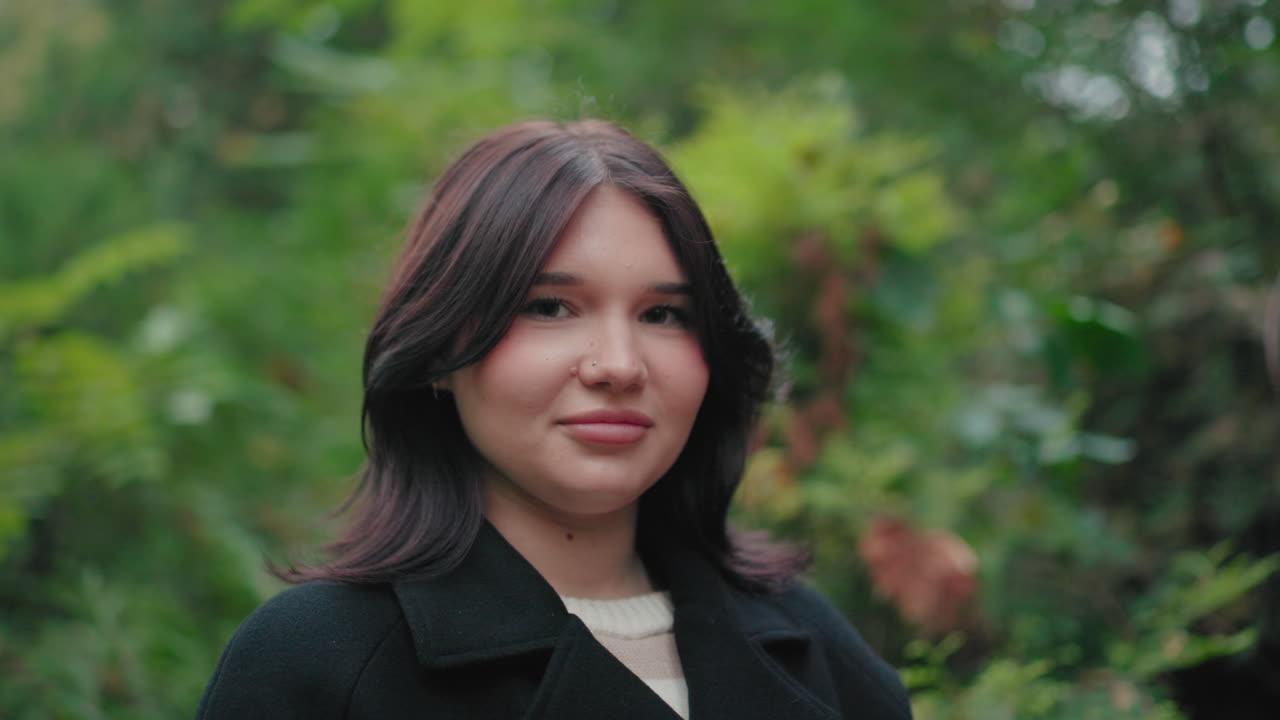 Portrait of stylish tourist standing before blur green autumn trees, smiling warmly in black coat; serene outdoor mood with soft bokeh background, youthful face framed by dark hair in forest setting