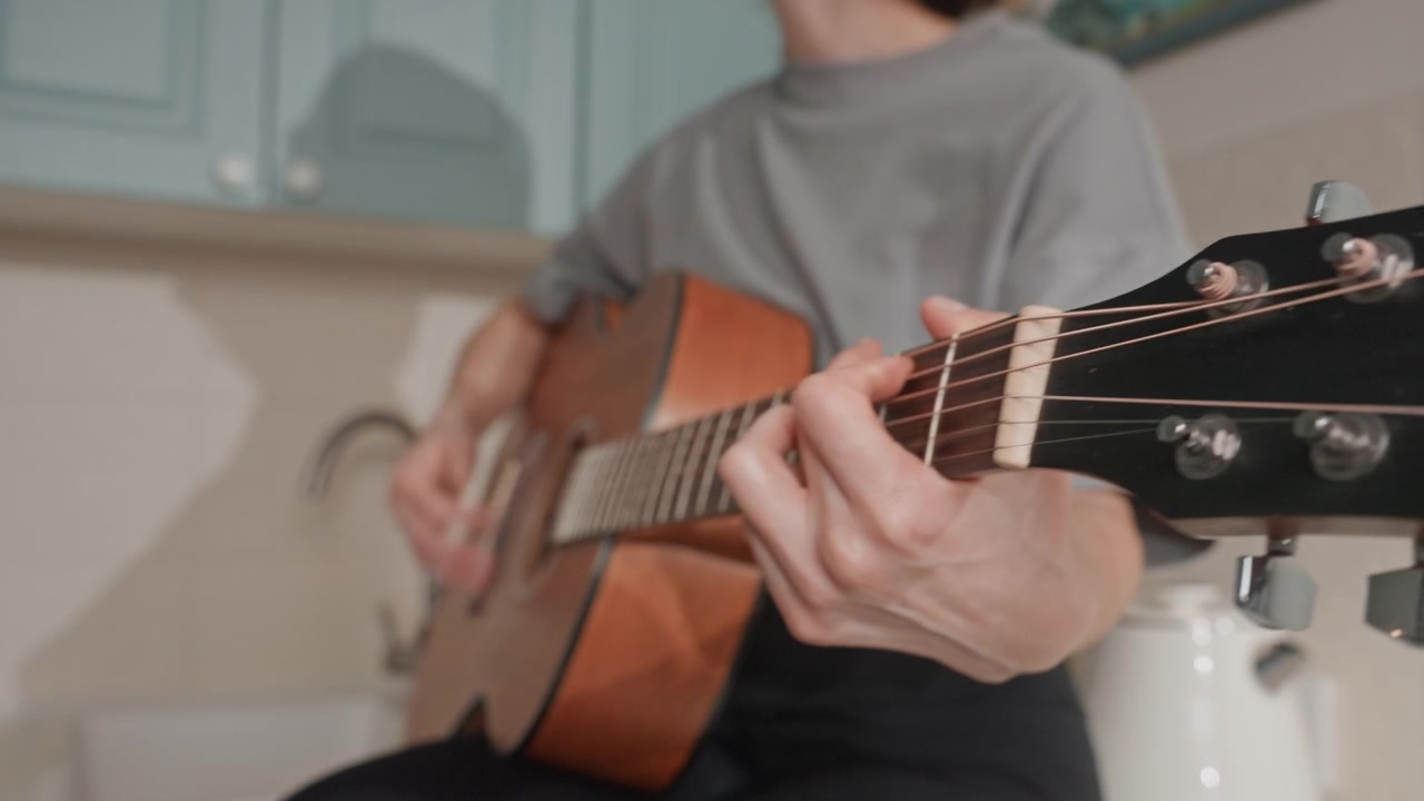 Closeup acoustic guitar in kitchen showing hands and fretboard, home songwriter gently fingerpicking slow chords, warm natural light, soft ambient mood, intimate expression of music lover and quiet