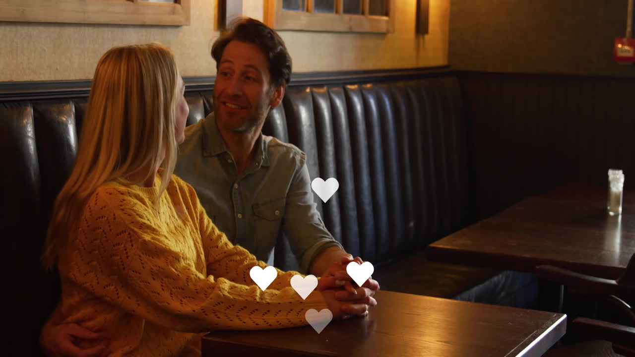 Hearts animation over couple holding hands and smiling in cozy restaurant