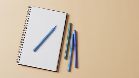 Overhead-view-of-blue-markers-with-notebook-with-copy-space-on-beige-background,-in-slow-motion