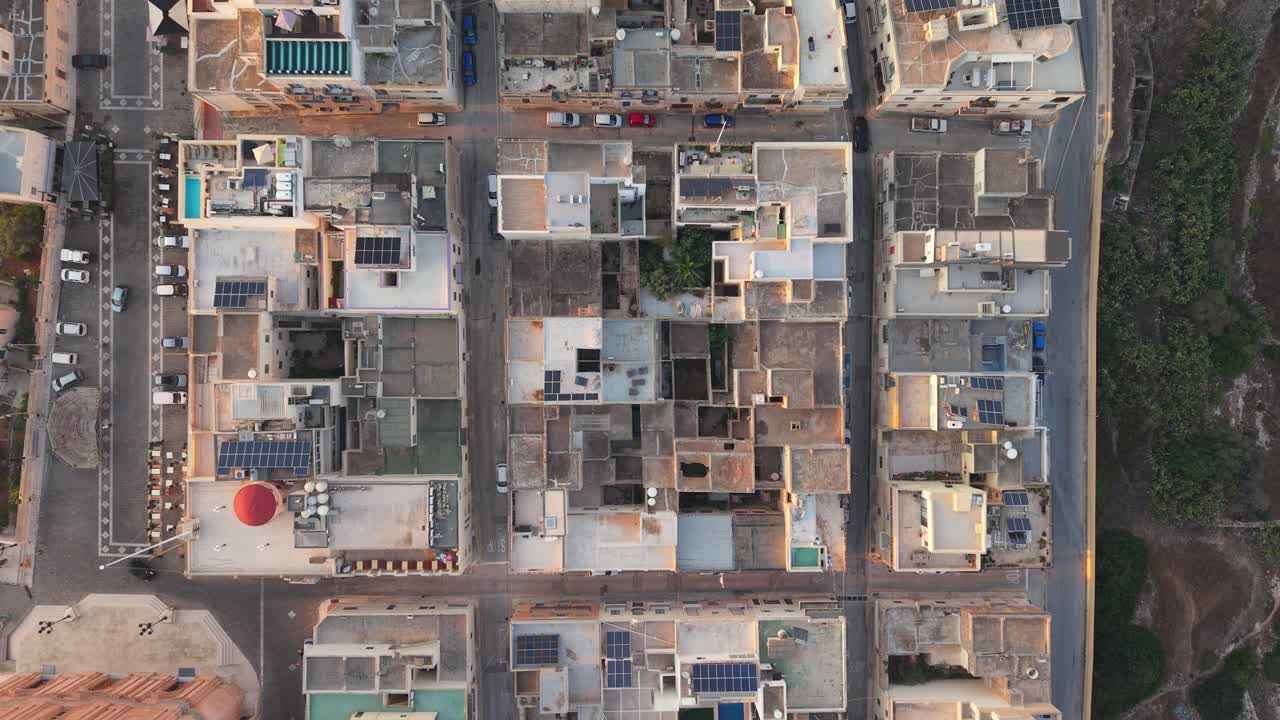 An overhead drone video of a Mediterranean city block showing flat rooftops, solar panels, narrow streets, and parked cars