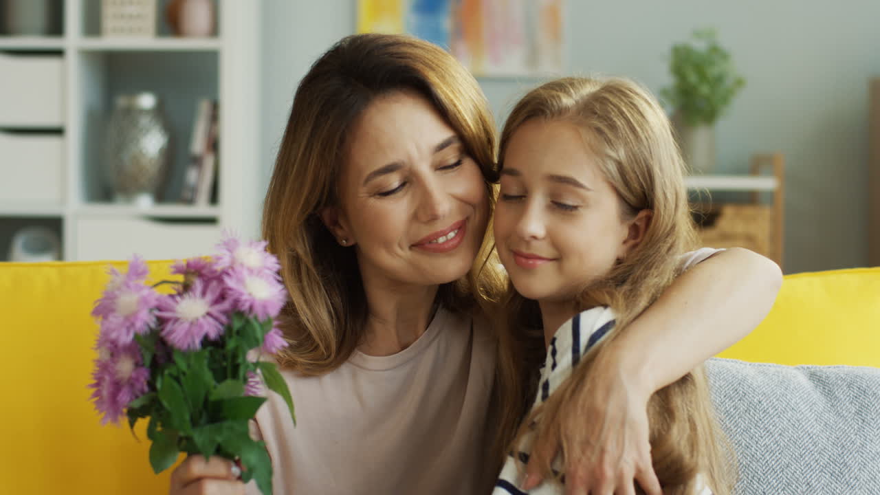 Close Up Of Teenager Girl Giving Flowers To Her Mother And Hugging Her On The Sofa In The Living Room