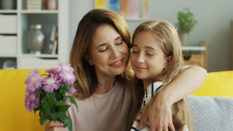 close up of teenager girl giving flowers to her mother and hugging her on the sofa in the living room