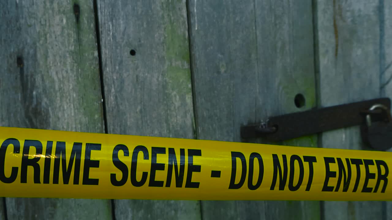 Video gliding over a yellow colored forensic police crime scene investigation tape ribbon line that is in front of a rusty brown and old emtallic padlock on a wooden farm door outside during day time