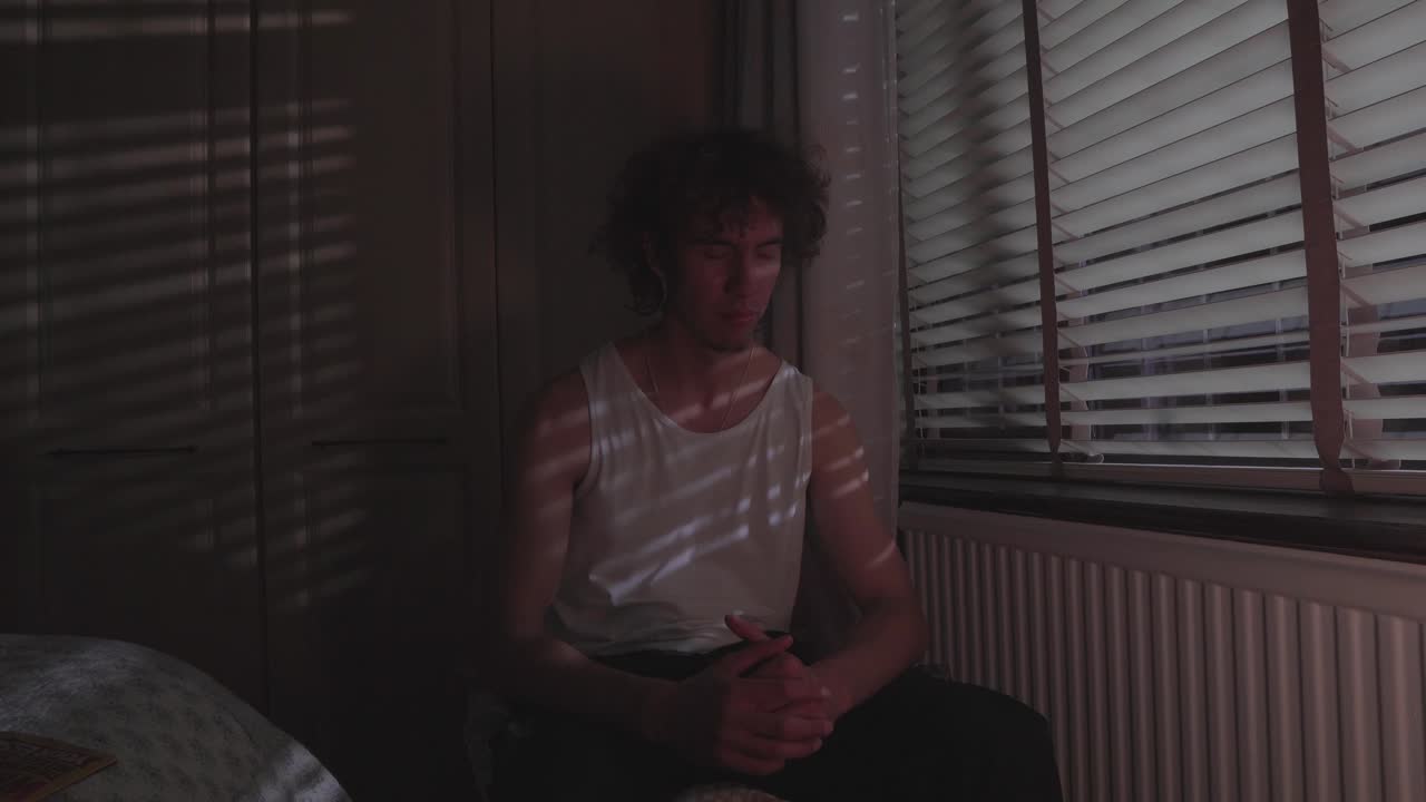 Man in emotional state sits hunched forward on bed in dim light with blinds casting lines