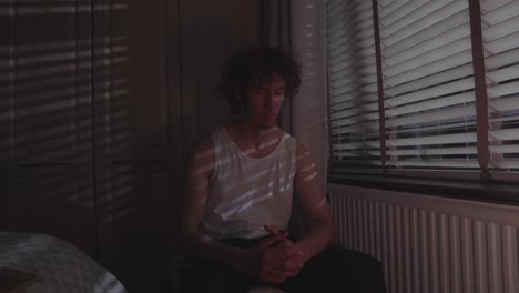 man in emotional state sits hunched forward on bed in dim light with blinds casting lines