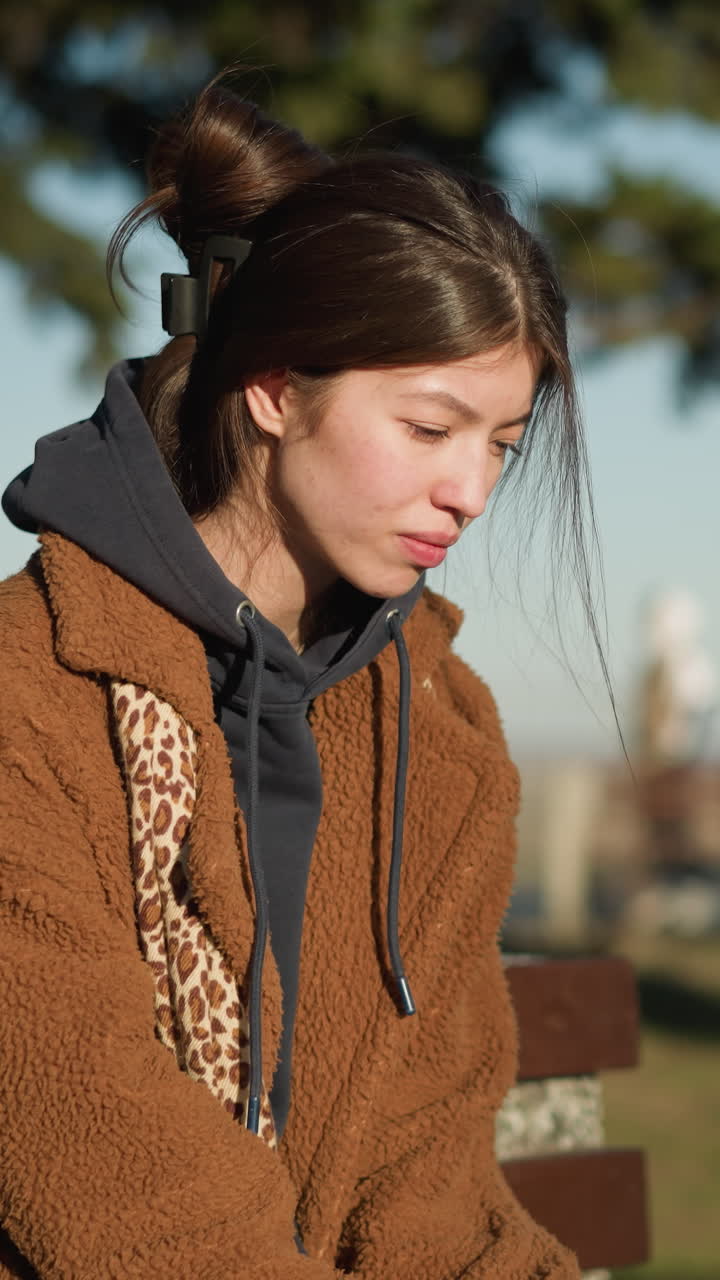 한 소녀가 도시 공원의 벤치에 혼자 앉아서 머리를 부드럽게 흔들면서 불행하고 생각에 빠져있는 것처럼 보입니다. 그녀는 갈색 코트, 파란 청바지, 색 신발을 입고 있습니다.