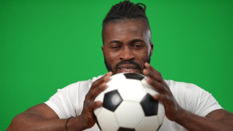 motivated confident african american sportsman squeezing football ball looking at camera. front view portrait of concentrated man posing at green screen chromakey background.