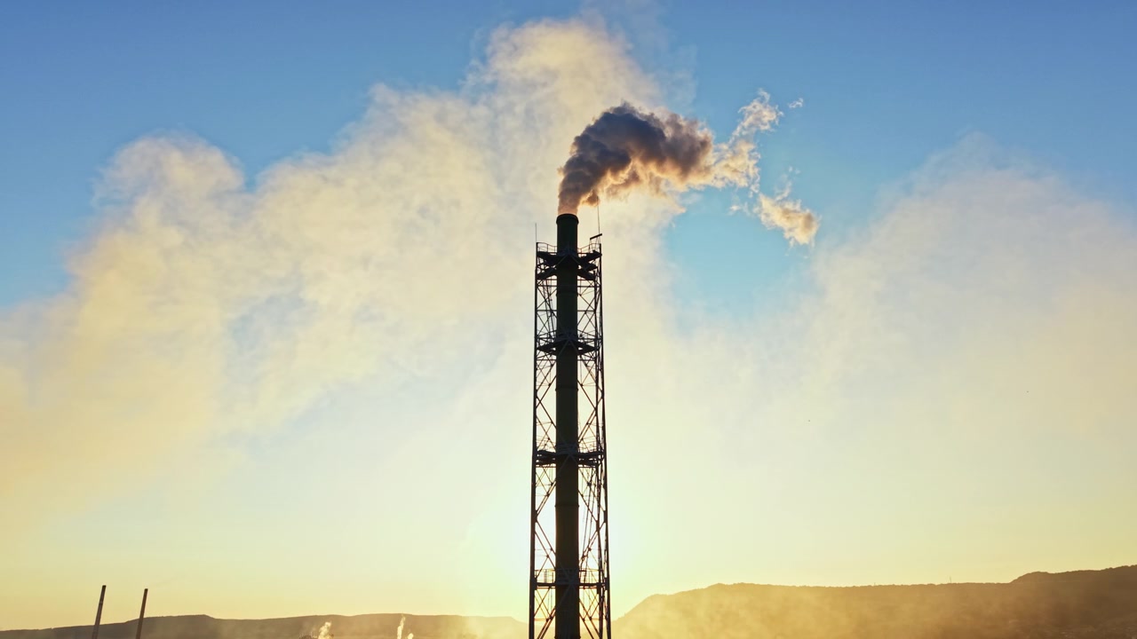 Factory smoke rises from a tall chimney during sunset creating a contrast with the sky and surrounding landscape