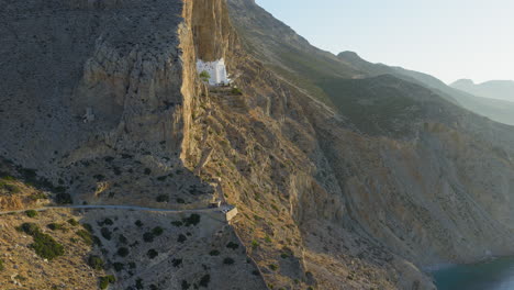 Sunrise light on Hozoviotissa Monastery against rocky cliffs in Amorgos, Greece, sideview aerial drone approach
