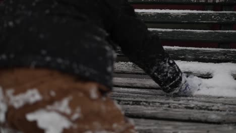 Young-boy-wearing-a-coat-playing-with-snow-outside-on-a-cold,-winter-day-in-December-with-a-snow-covered-bench-during-Christmas-break-in-a-small-town-in-the-midwest