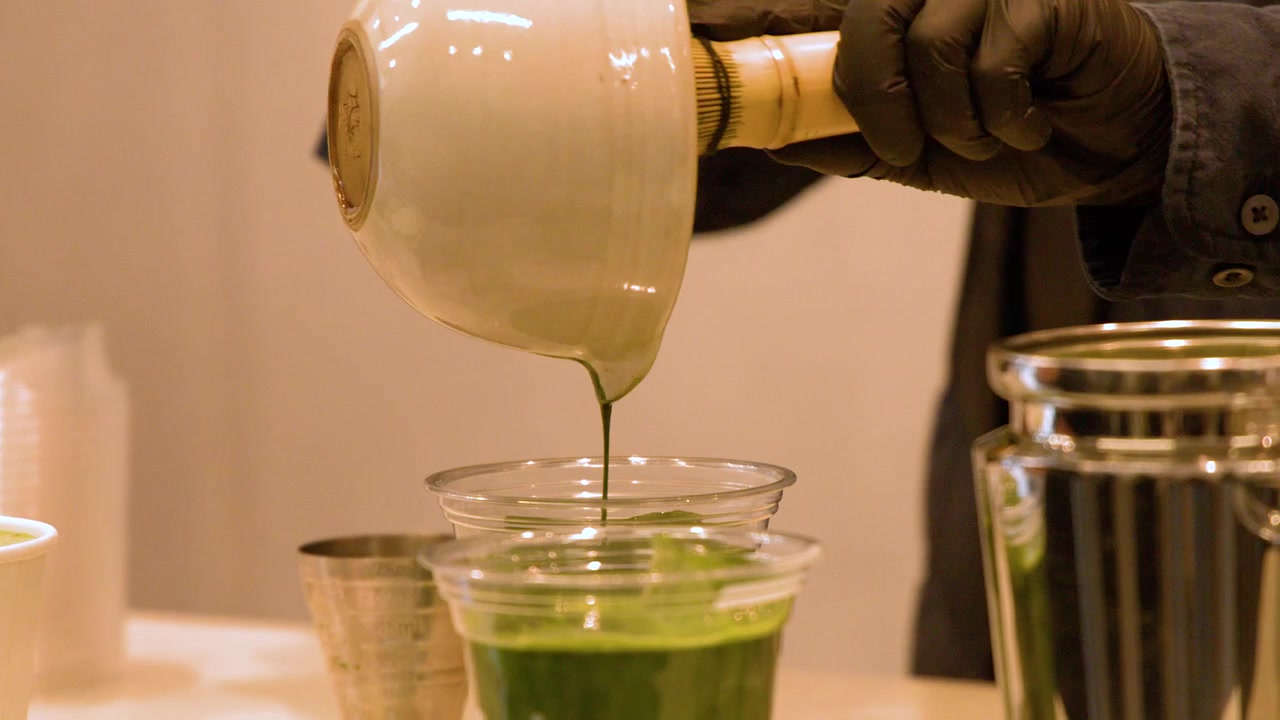 Primo piano di un barista che versa tè matcha verde brillante da una ciotola di ceramica in tazze.