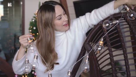 mujer feliz colgando luces navideñas en una silla en una sala de estar con adornos navideños y árbol de navidad