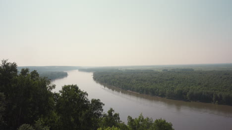 serene panoramic view of wide calm river winding through lush green countryside under pale hazy sky, surrounded by dense tree line and soft morning light in peaceful natural landscape
