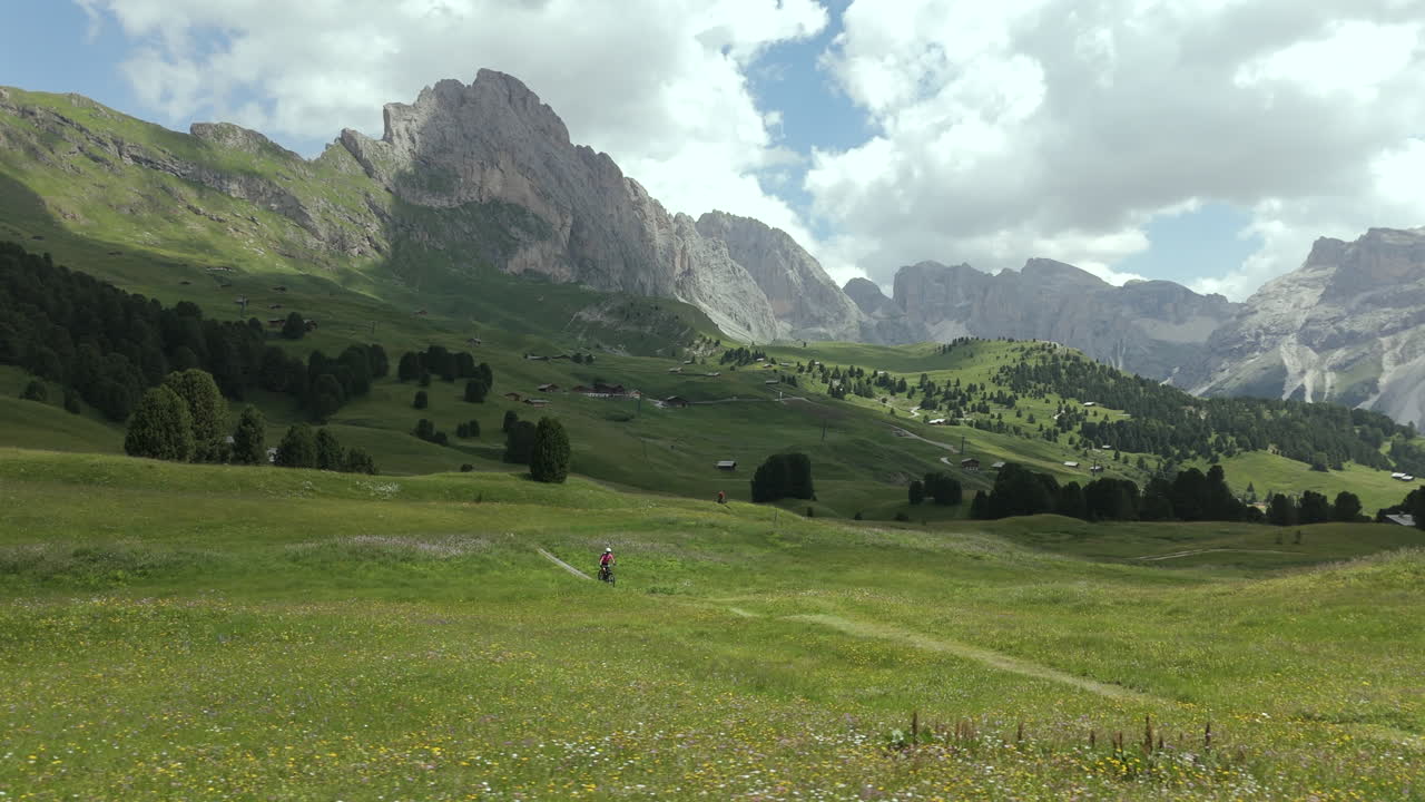 한 자전거 타는 사람 이 이탈리아 의 돌로미트 산맥 의 바위 산꼭대기 들 을 환상적 으로 볼 수 있는 푸른 초원 들 을 가로질러 달린다.