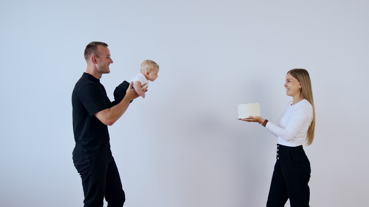 Man in black clothes walks holding his infant son. Woman with cake in hands approaches her husband and son. White backdrop.