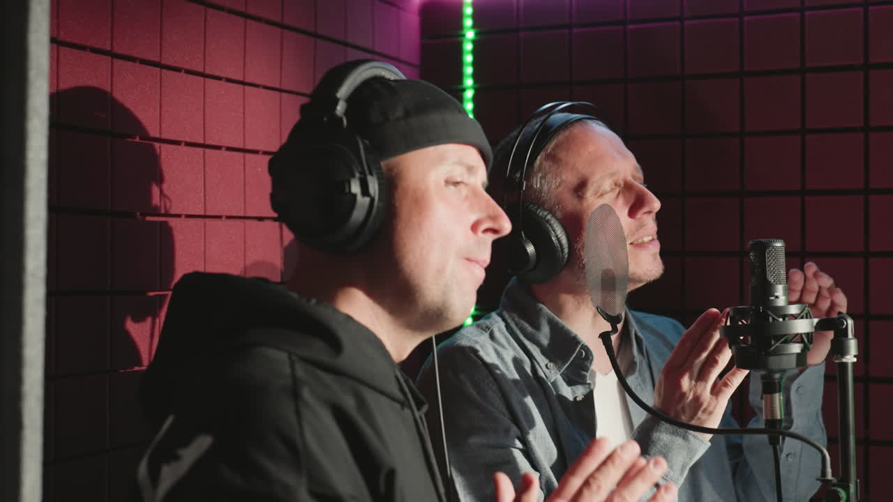 두 남자가 헤드폰을 착용하고, 한 사람은 검은 모자와 후디를 입고, 다른 한 사람은 파란 셔츠와  셔츠를 입고 소음 방지 녹음 스튜디오에서 손을 박고 있습니다.