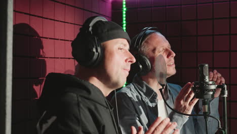 two men wearing headphones, one in a black cap and hoodie and the other in a blue shirt and white undershirt, are clapping hands in a soundproof recording studio. they are mid-performance