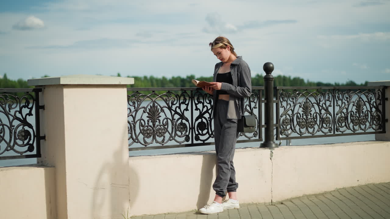 дама в серой одежде и черной сумочке стоит рядом с забором, читает книгу с сосредоточенным вниманием, когда она потирает нос, деревья видны на расстоянии, и фонарь находится поблизости