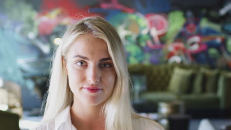 young white woman with blonde hair in front of mural in workplace smiling to camera, close up