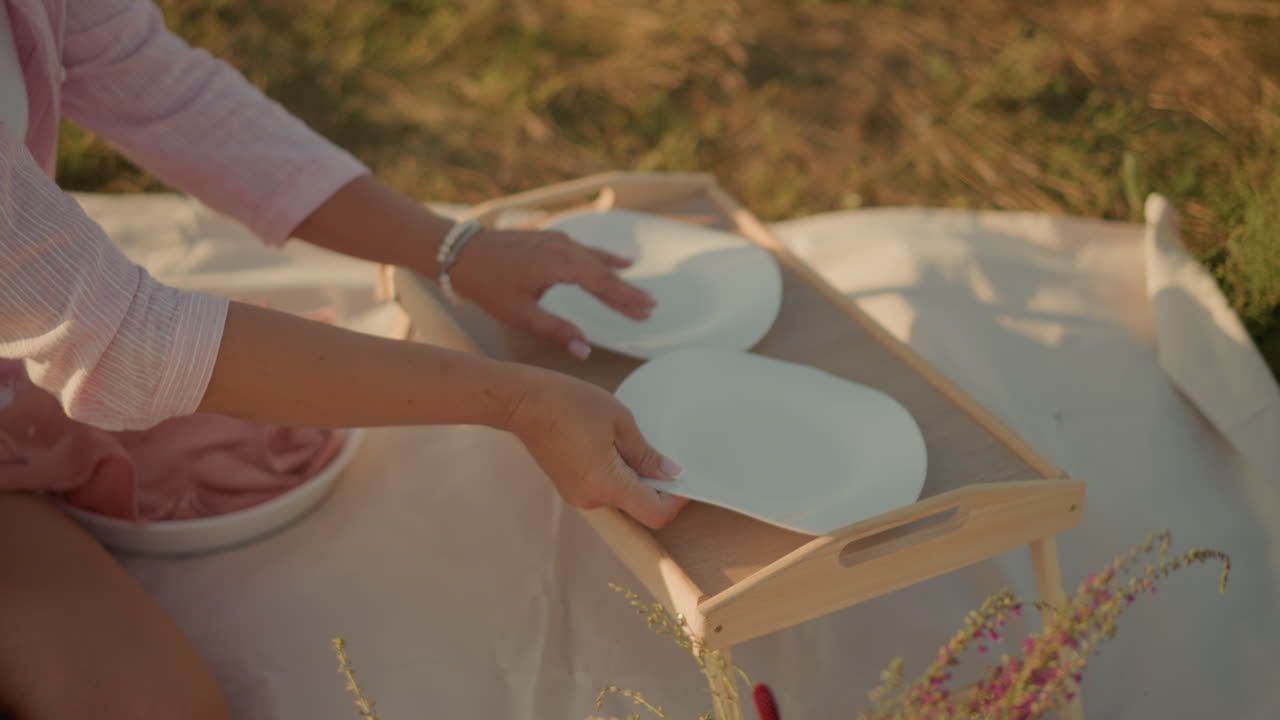 에 인 바구니에 꽃이 있는 야외 매트에 앉아 있는 여자, 빛 아래 피크닉 테이블에 접시를 배열하고, 손으로 접시를 조정하며, 따뜻한 날에 평화롭고 이한 피크닉 분위기를 만니다.