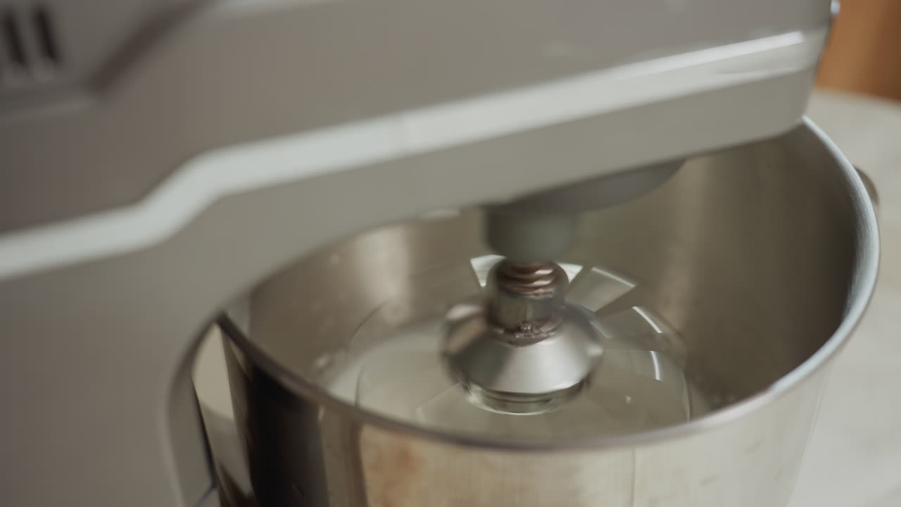 Close up of electric mixer blending mashed white ingredient inside stainless steel bowl on kitchen table, with wooden baking stand nearby, showing rapid mixing action in warm indoor setting