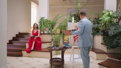 un artista dibuja a una mujer con ropa roja en una galería. una hermosa dama con un vestido carmesí posa para un retratista experto en una cabaña decorada con plantas exóticas.