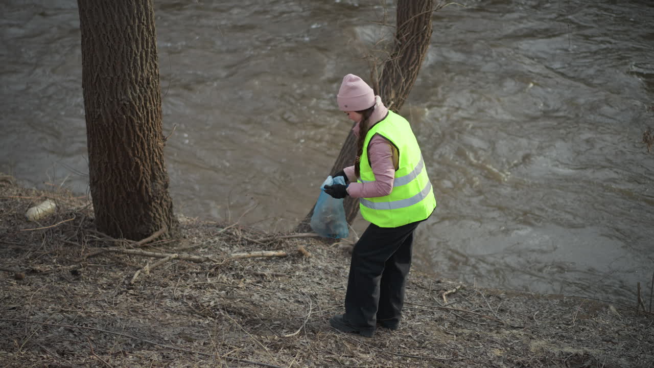 Woman in high visibility vest and pink beanie collects litter in blue plastic bag along muddy riverbank during environmental cleanup, bending to pick up trash near fast-flowing water on overcast day