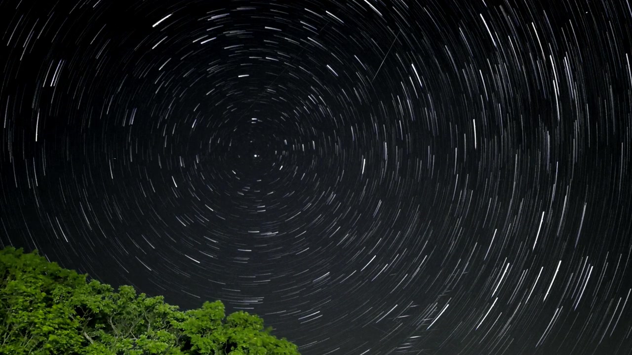 Long exposure star trails circling across night sky above Cairngorms, capturing Earth's rotation, bright stars, and silhouetted treetops beneath clear night conditions