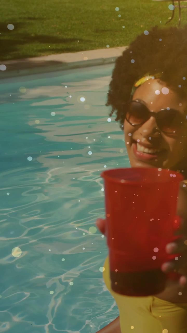 Vertical video: Woman sitting poolside raising red cup toward camera causing animated bokeh for ads