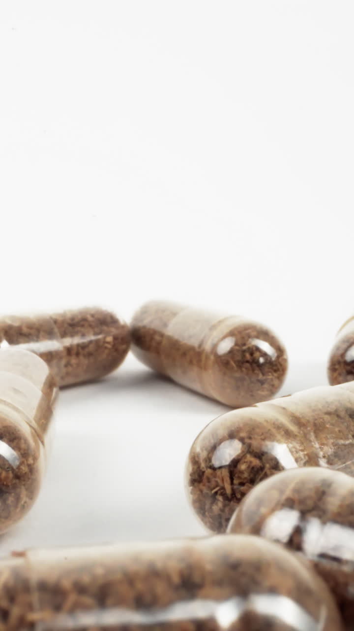 primer plano de suplementos herbáceos marrones en cápsulas transparentes esparcidas en blanco