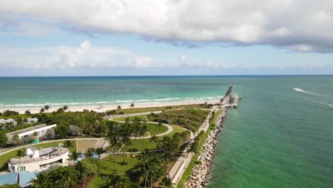 Flyover-of-pier-and-beach-along-a-jetti-with-a-park-by-the-seaside-coast-and-waves-crashing-on-the-sand