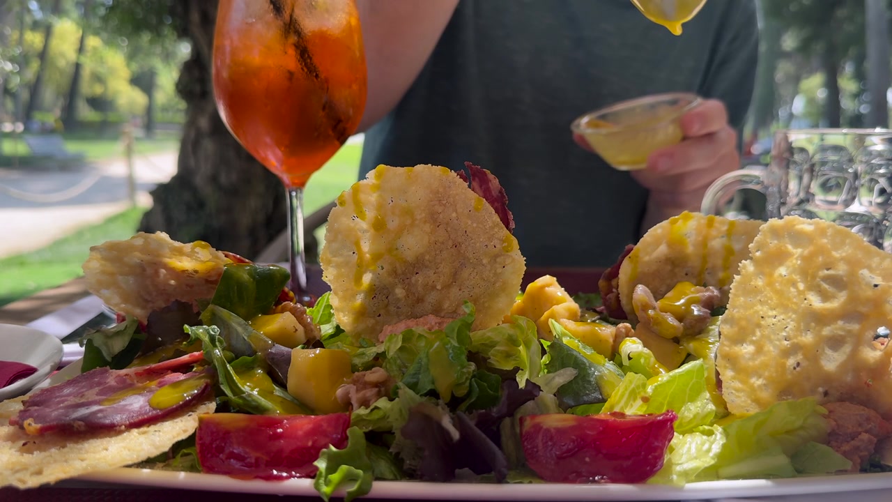 Primo piano della mano e della parte superiore del corpo di un giovane uomo caucasico che indossa un maglione scuro, mentre condisce un'insalata mediterranea colorata con salsa al mango, seduto all'aperto su una terrazza