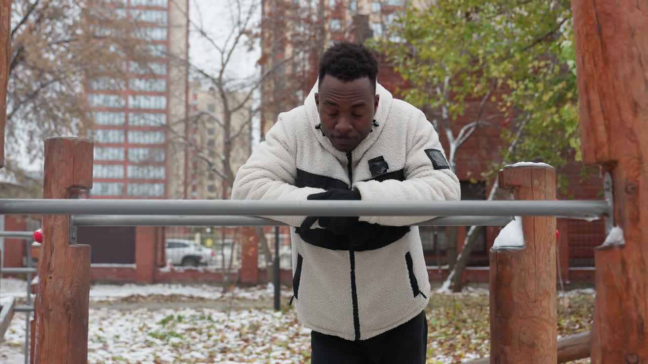 joven negro se apoya en una barra de hierro, descansando después del entrenamiento, mira a su alrededor pensativo en un gimnasio al aire libre con el suelo cubierto de nieve, árboles y edificios residenciales en el fondo