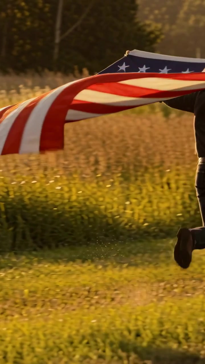 Video vertikal: Angin mengibarkan bendera Amerika, orang dewasa memegangnya dan melintasi ladang hangat mengenakan mantel gelap.