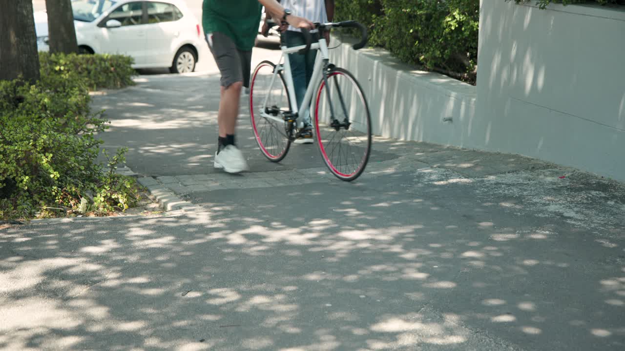 Caminhantes diversos em uma calçada sombria, um ciclista descendo e empurrando sua bicicleta de estrada, no trajeto diário.
