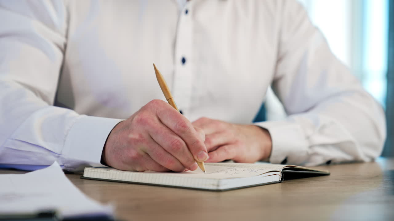 Hands of a male wearing white shirt taking notes into the paper book. Close up view of a handwriting with a ball pen.