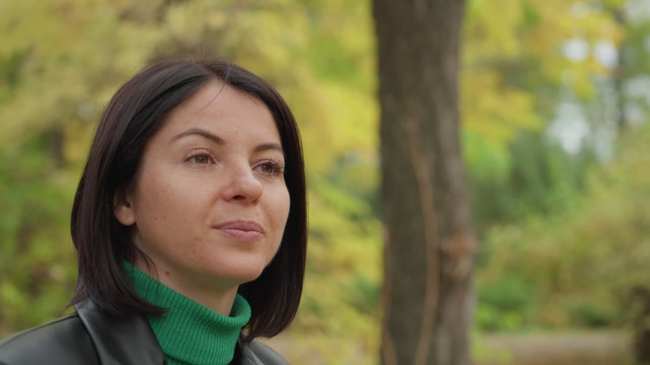 Donna caucasica con giacca di pelle che riflette silenziosamente in un parco autunnale, indossa un dolcevita verde, bokeh delicato di alberi e fogliame, dettagli ravvicinati del viso, sorriso gentile e sguardo pensieroso, caldo naturale