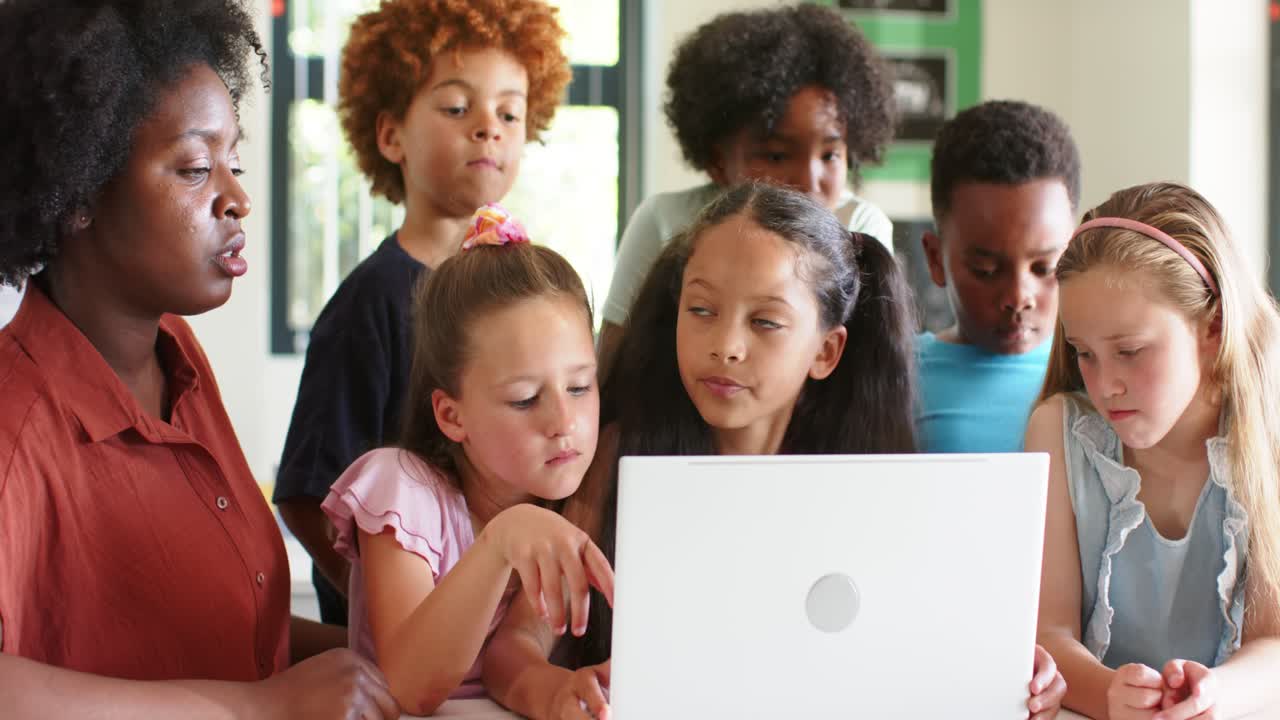 African American teacher in rust top pointing at white laptop, teaching children at classroom table