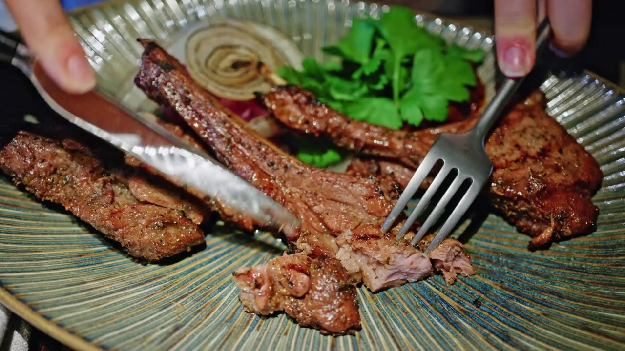 Gambar close-up iga babi panggang yang dilumuri bumbu gurih, sedang dipotong dengan pisau dan garpu di atas piring keramik