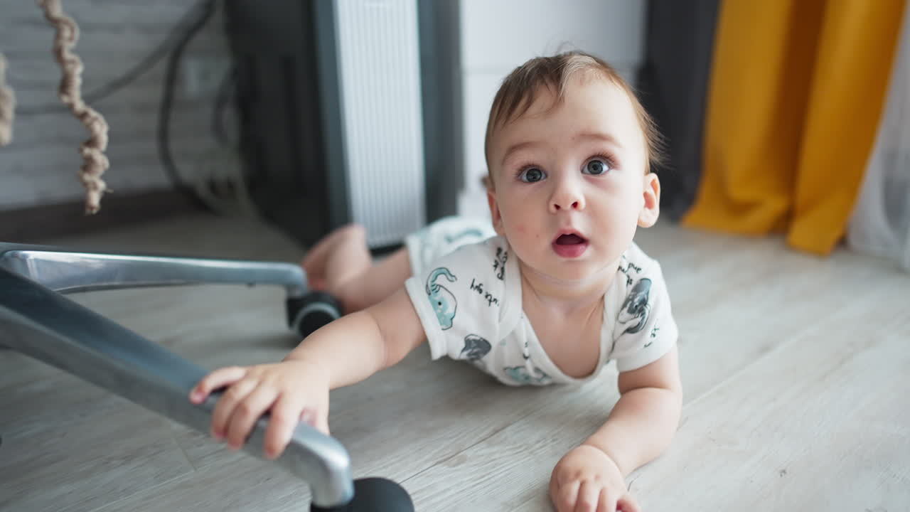 Adorable baby crawling by the floor, touching the wheels of chair. Camera approaches cute boy quickly and he touches camera with curiosity. Close up.