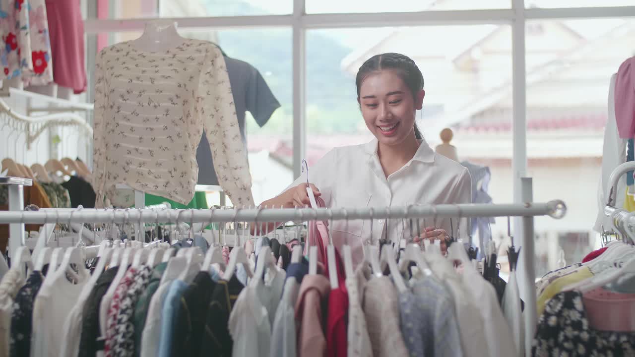 옷 가게 의 아시아인 여성 소유자 가 가게 에서 재고 를 확인 하는 동안 레일 에 드레스 를 매달고 있다