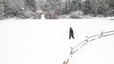 A-lone-woman-walks-past-a-fence-and-a-snowman-across-a-snow-covered-park-field,-aerial-orbit