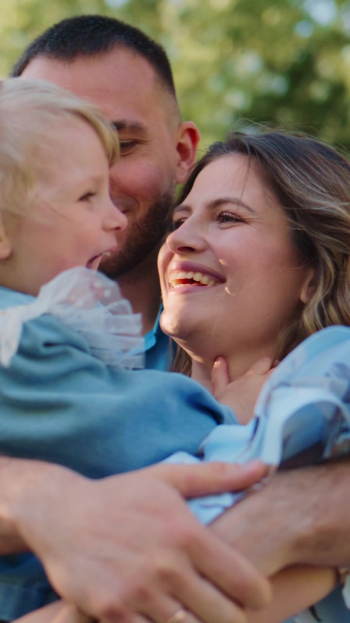 Una giovane famiglia condivide un momento gioioso all'aperto. Il padre tiene in braccio il bambino, la madre sorride dolcemente. Il bambino si avvicina, giocoso e curioso. L'ambiente caldo e illuminato dal sole evoca il legame familiare. Ideale per genitorialità, lifestyle.