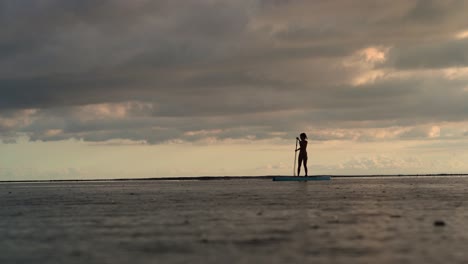 silhouette of girl on stand up paddle moving on ocean during sunset and rain