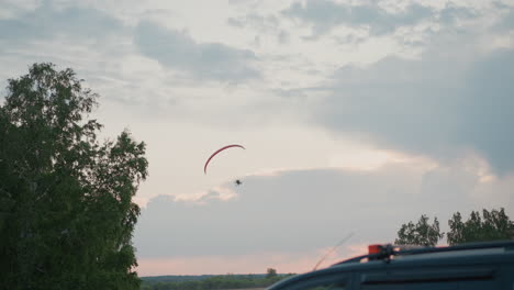 close up of parked suv near grove of birches with powered paraglider launching overhead at dusk under pastel sky, capturing serene countryside adventure and thrill of flight over open grass