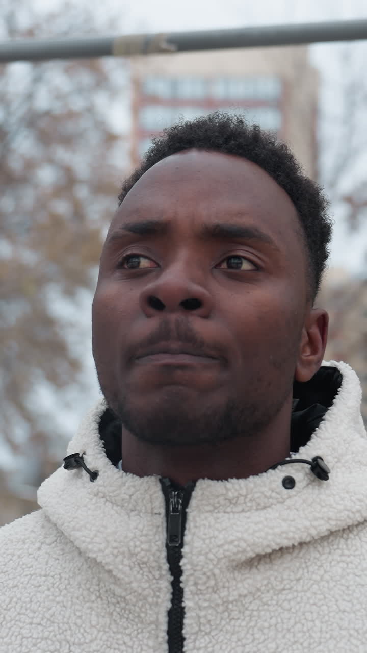 joven decidido que parece centrado mientras está de pie en un área de entrenamiento al aire libre, con una cálida chaqueta de lana, parece profundamente pensativo, el fondo presenta árboles y un entorno urbano