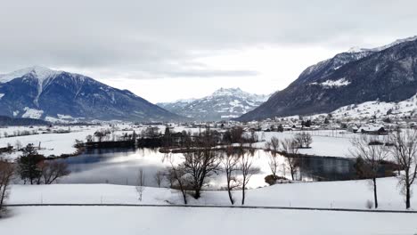 Vuelo-Aéreo-Hacia-Adelante-Sobre-Una-Carretera-Nevada-Con-Tráfico-Y-Un-Lago-En-Los-Alpes-Suizos-Durante-El-Día-Nublado---Pequeña-Ciudad-Al-Fondo