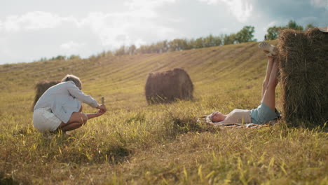 photographer capturing her sister photo outdoors as she places her legs on hay in vast farmland, sunny day, relaxed countryside atmosphere, creativity in nature