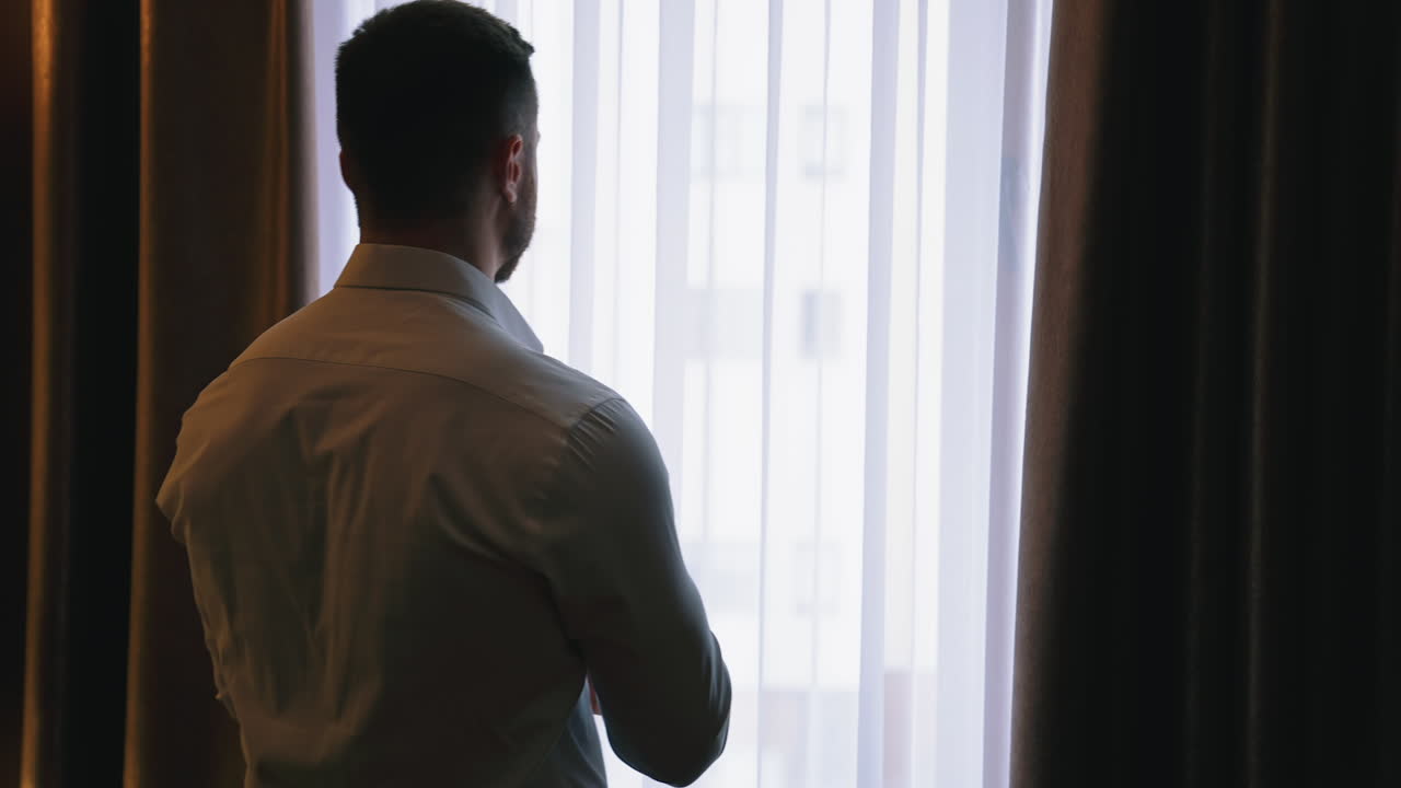 Man in hotel near the window. Young businessman in white shirt standing near the window and viewing through it in the hotel room. Back view.